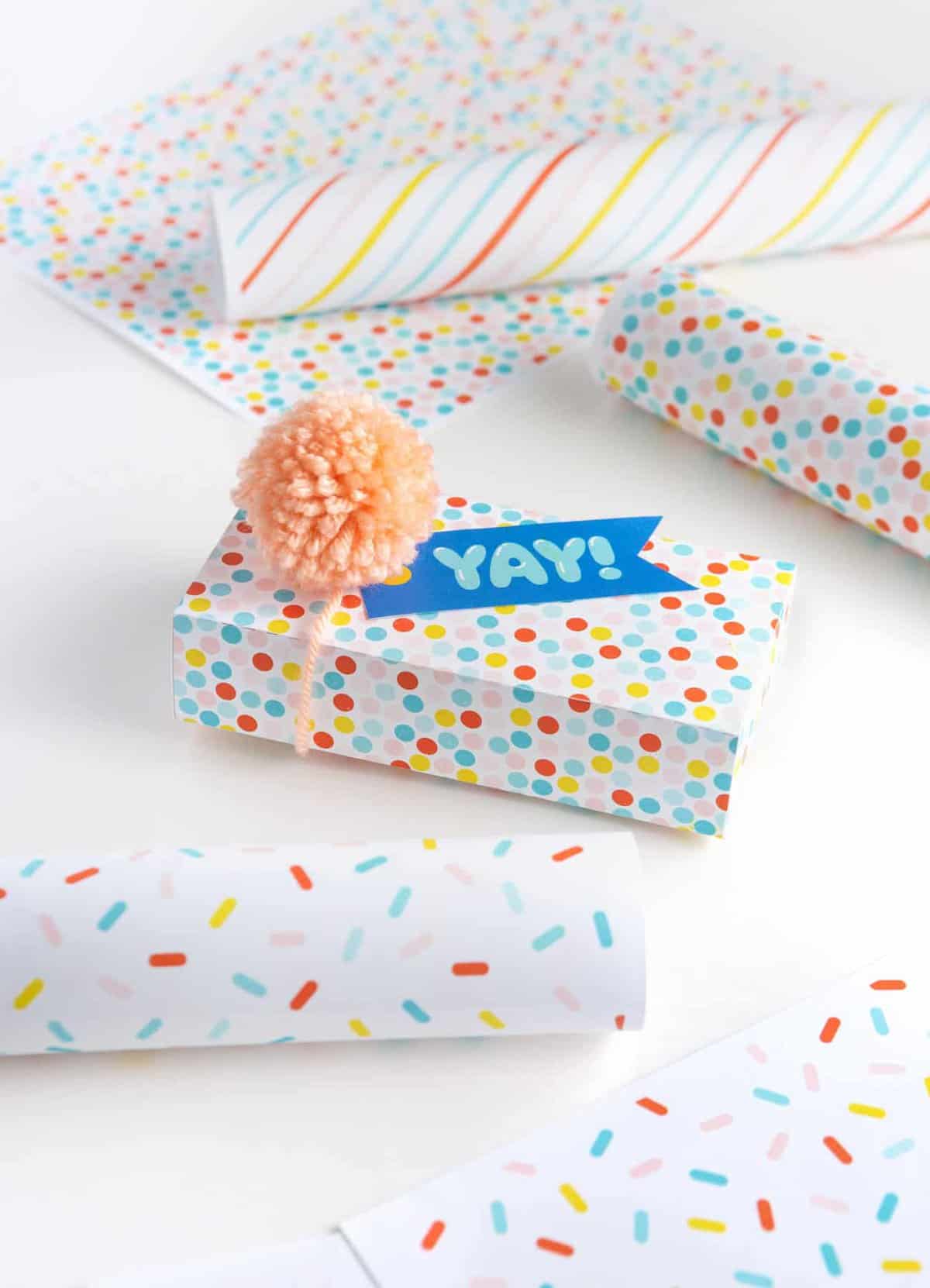 Free printable birthday wrapping paper gift box with blue yay tag and pink pom pom. Patterns include rainbow polka dot, rainbow sprinkle, and rainbow stripe.