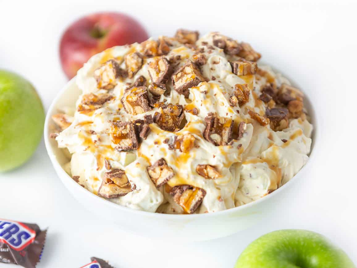 Bowl of creamy apple snickers salad dessert, topped with chopped snickers candy bars, apples, and caramel sauce