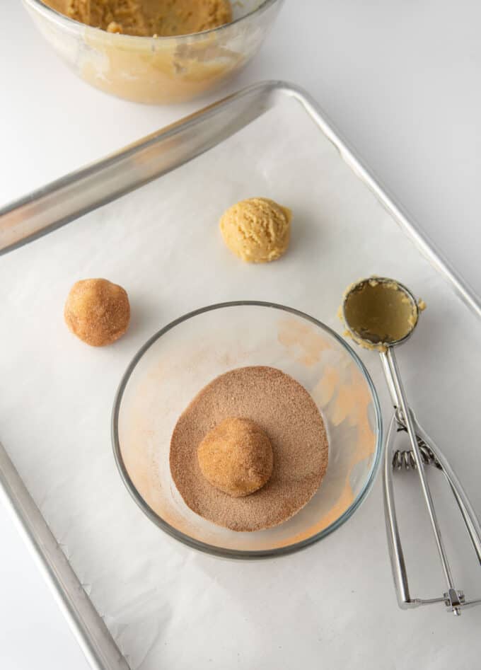 Metal baking pan with scoops of cookie dough and bowl of cinnamon sugar to roll cookies in