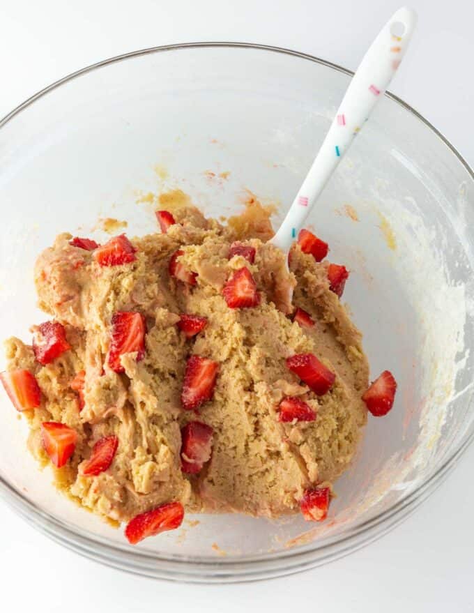 Cookie dough, sliced strawberries and a confetti spatula are in a glass bowl on top of a white countertop