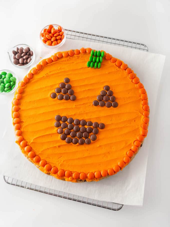 round chocolate chip cookie cake with orange frosting and m&m's pressed on top to create a smiling Halloween jack-o-lantern pumpkin face