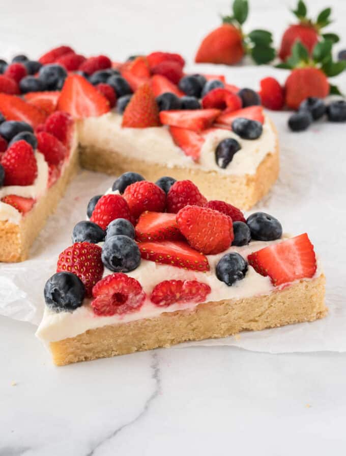 slice of sugar cookie berry pizza with cream cheese frosting and sliced strawberries, raspberries, and blueberries
