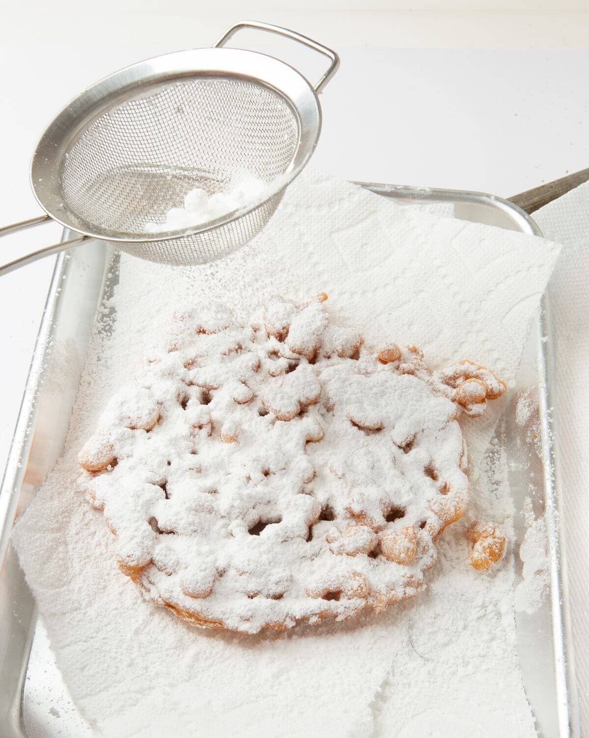 funnel cake on paper towel lined tray being sprinkled with powdered sugar using mesh colander