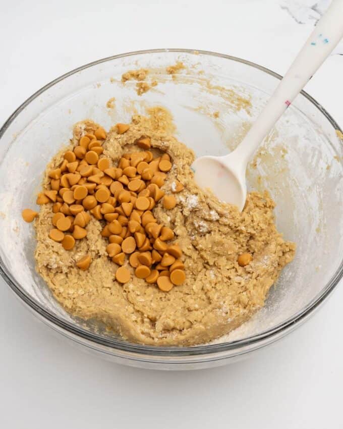 adding butterscotch chips to oatmeal scotchie dough in glass mixing bowl with rubber spatula
