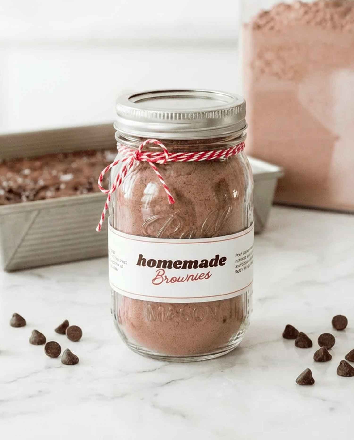 Homemade brownie mix in a mason jar tied with red and white twine, with a printable label and chocolate chips scattered on a white marble surface