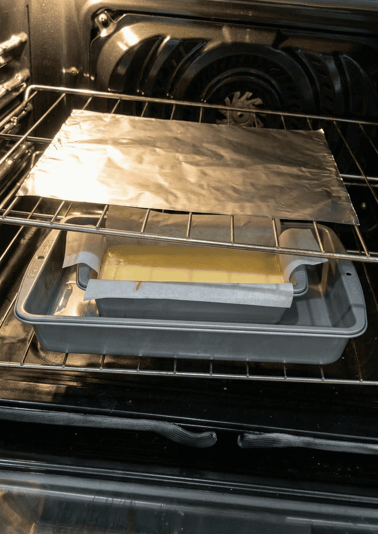 A loaf pan of cheesecake batter in a water bath inside a preheated oven, with a loose sheet of aluminum foil tented on the rack above to prevent overbrowning