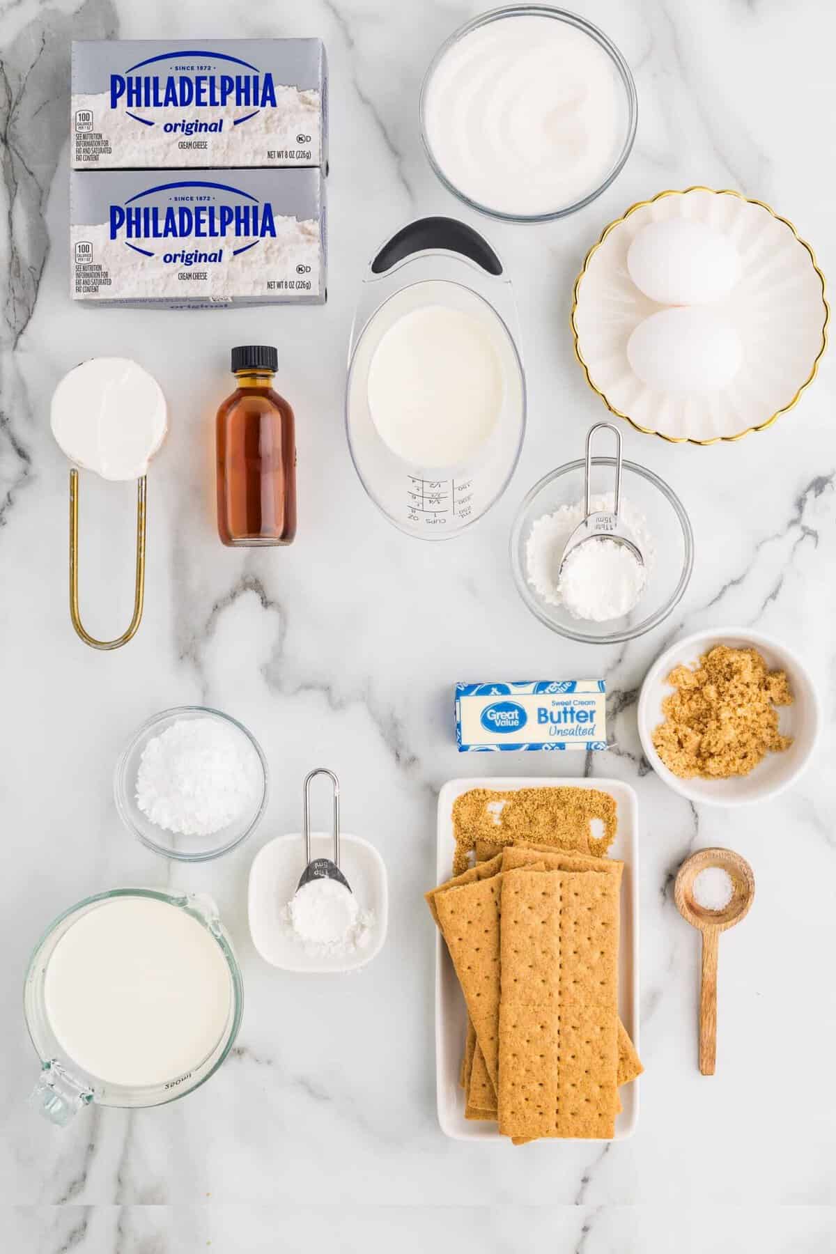 Flat-lay of loaf pan cheesecake ingredients on a marble surface, including Philadelphia cream cheese blocks, sour cream, eggs, vanilla extract, heavy cream, sugar, graham crackers, butter, and cornstarch