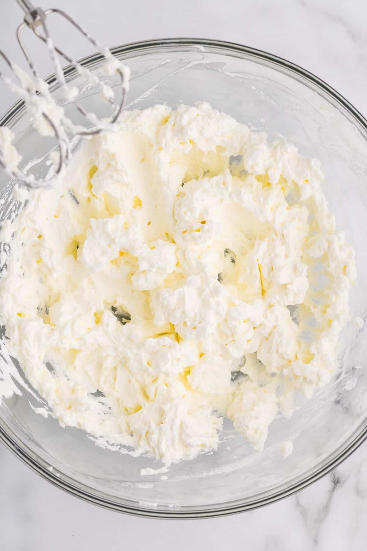 Overhead view of cream cheese being beaten with a hand mixer in a glass bowl, showing a fluffy and whipped texture at the start of making cheesecake filling