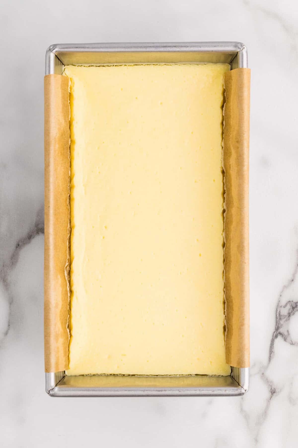 Overhead view of a fully baked cheesecake in a parchment-lined loaf pan, showing a smooth, pale yellow surface after baking