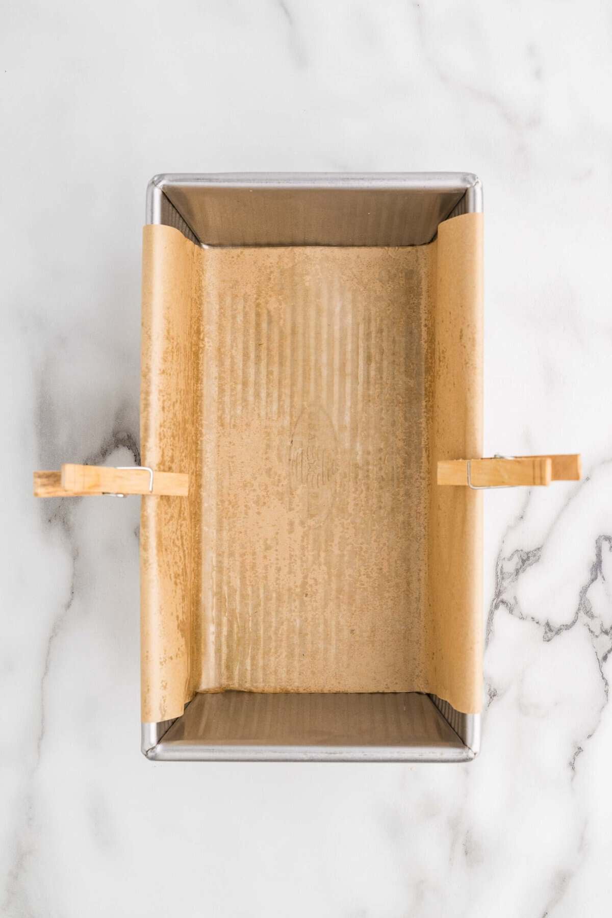 Overhead view of a loaf pan lined with parchment paper and secured with wooden clothespins, sitting on a marble surface
