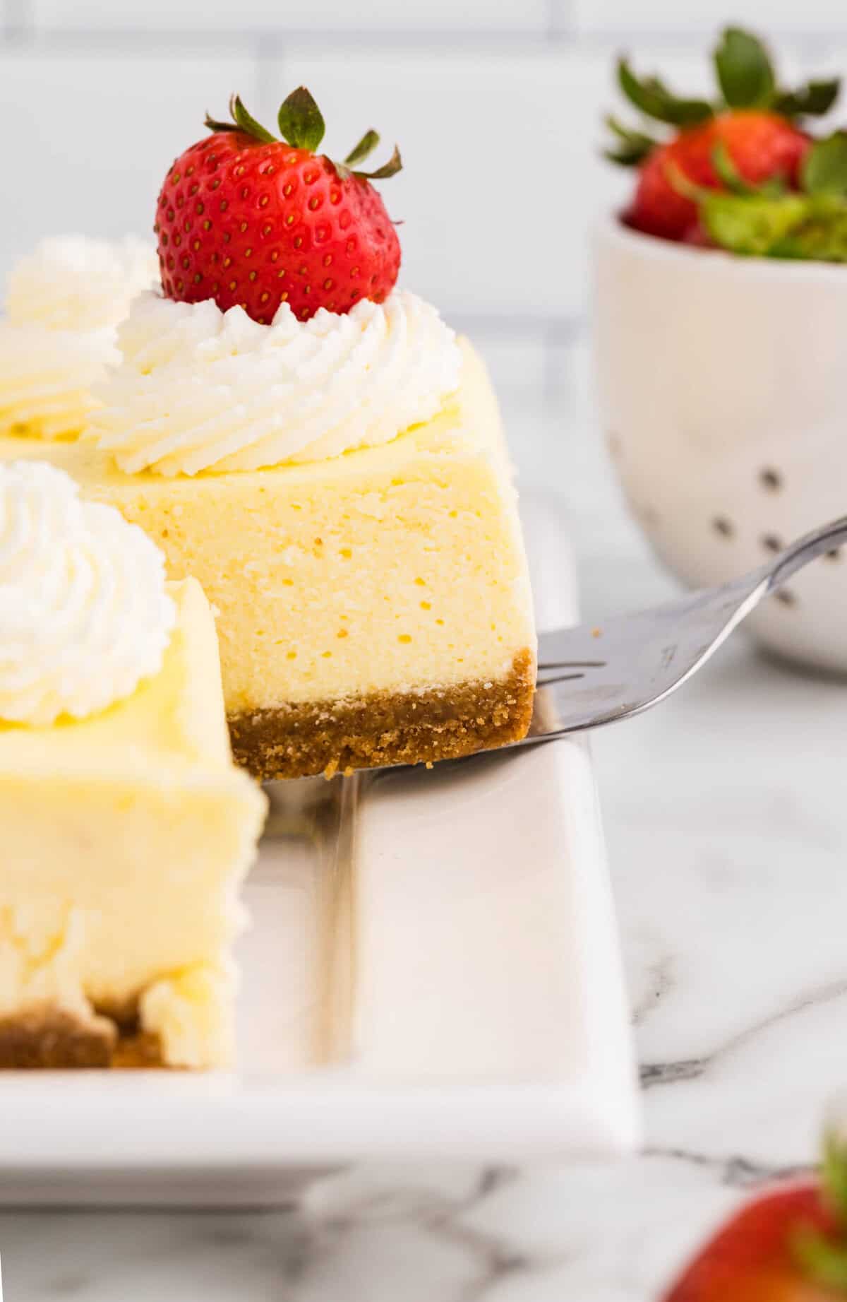 Close-up side view of a triangular loaf pan cheesecake slice topped with whipped cream and a fresh strawberry being lifted with a cake server, showing the smooth creamy filling and graham cracker crust