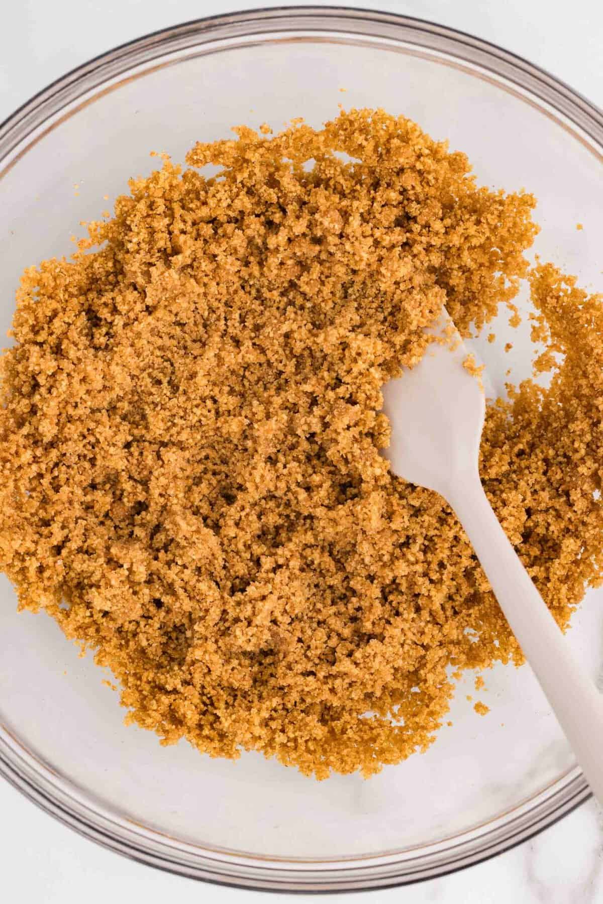 Overhead view of graham cracker crumbs mixed with melted butter in a glass mixing bowl, stirred with a white spoon to make cheesecake crust
