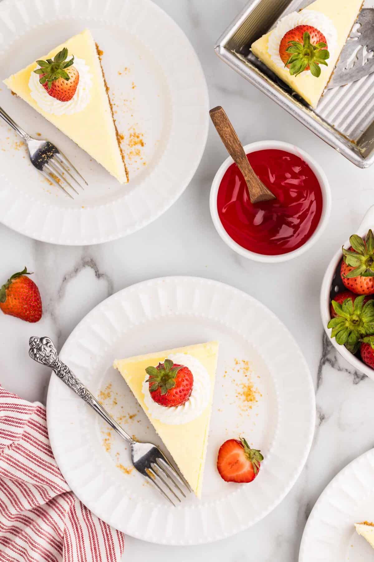 Overhead view of two white plates each with a triangular cheesecake slice topped with whipped cream and a strawberry, beside a bowl of strawberry sauce, fresh strawberries, and the remaining cheesecake in the loaf pan