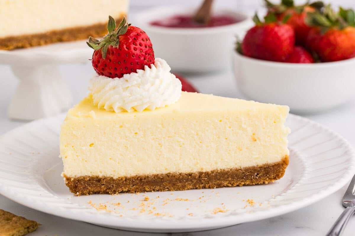 Close-up of a creamy cheesecake slice with graham cracker crust, topped with whipped cream and a strawberry on a white plate, with the full cheesecake on a cake stand and fresh strawberries in a bowl in the background