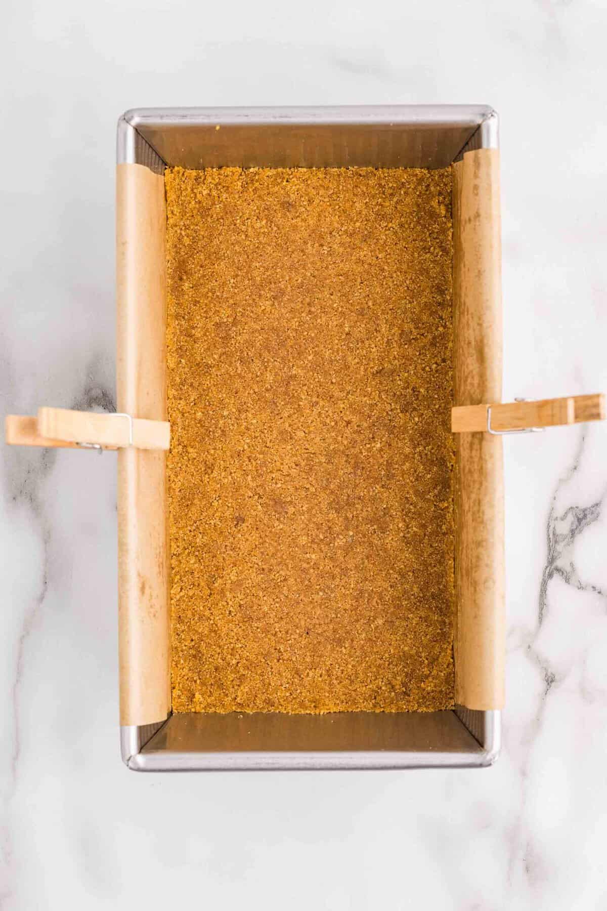 Overhead view of graham cracker crust pressed evenly into the bottom of a parchment-lined loaf pan secured with clothespins on a marble surface