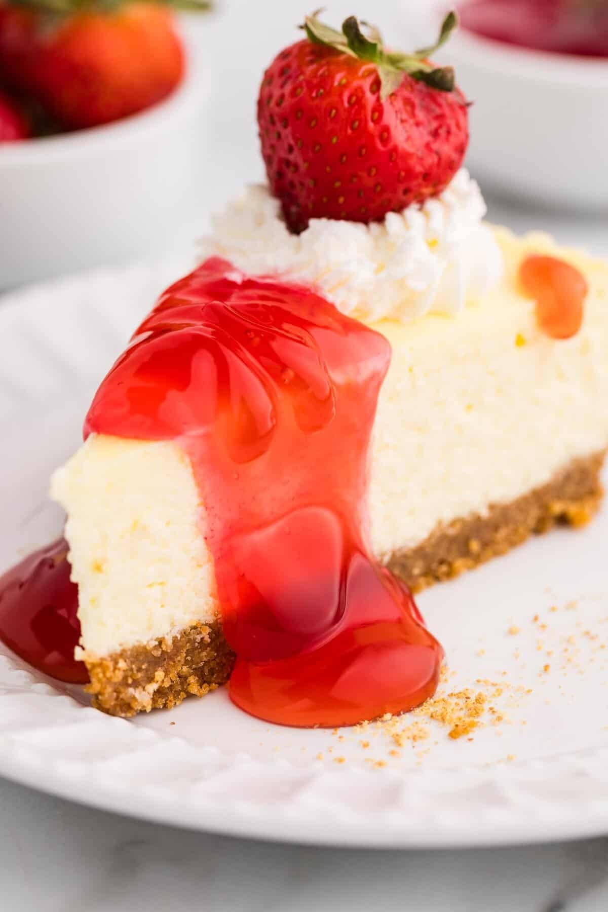 Close-up of a creamy cheesecake slice drizzled with strawberry sauce, topped with whipped cream and a fresh strawberry, served on a white plate