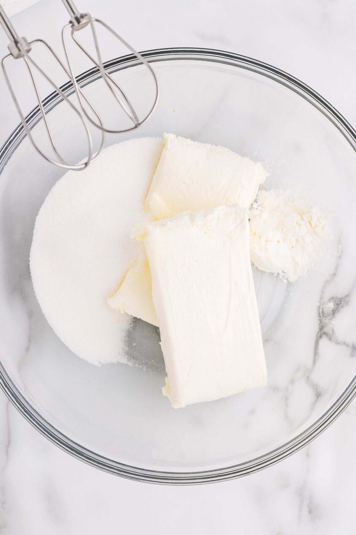 Overhead view of cream cheese blocks and granulated sugar in a glass mixing bowl with a hand mixer, ready to beat together for cheesecake filling