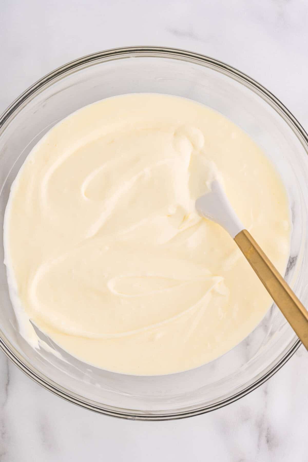 Overhead view of smooth, creamy cheesecake batter in a glass mixing bowl with a gold-handled spatula, ready to be poured into the pan