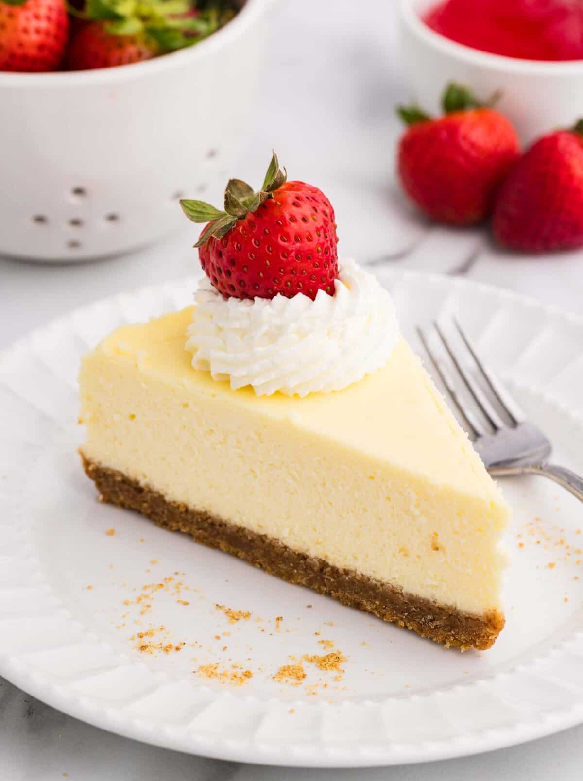 A slice of creamy loaf pan cheesecake with a graham cracker crust, topped with whipped cream and a fresh strawberry, served on a white plate with a fork and strawberries in the background