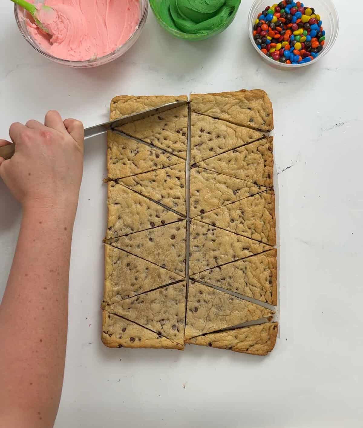 Overhead view of cookie cake cutting supplies: bowls of pink and green frosting, M&M seeds, and a rectangular cookie cake being cut into triangular slices