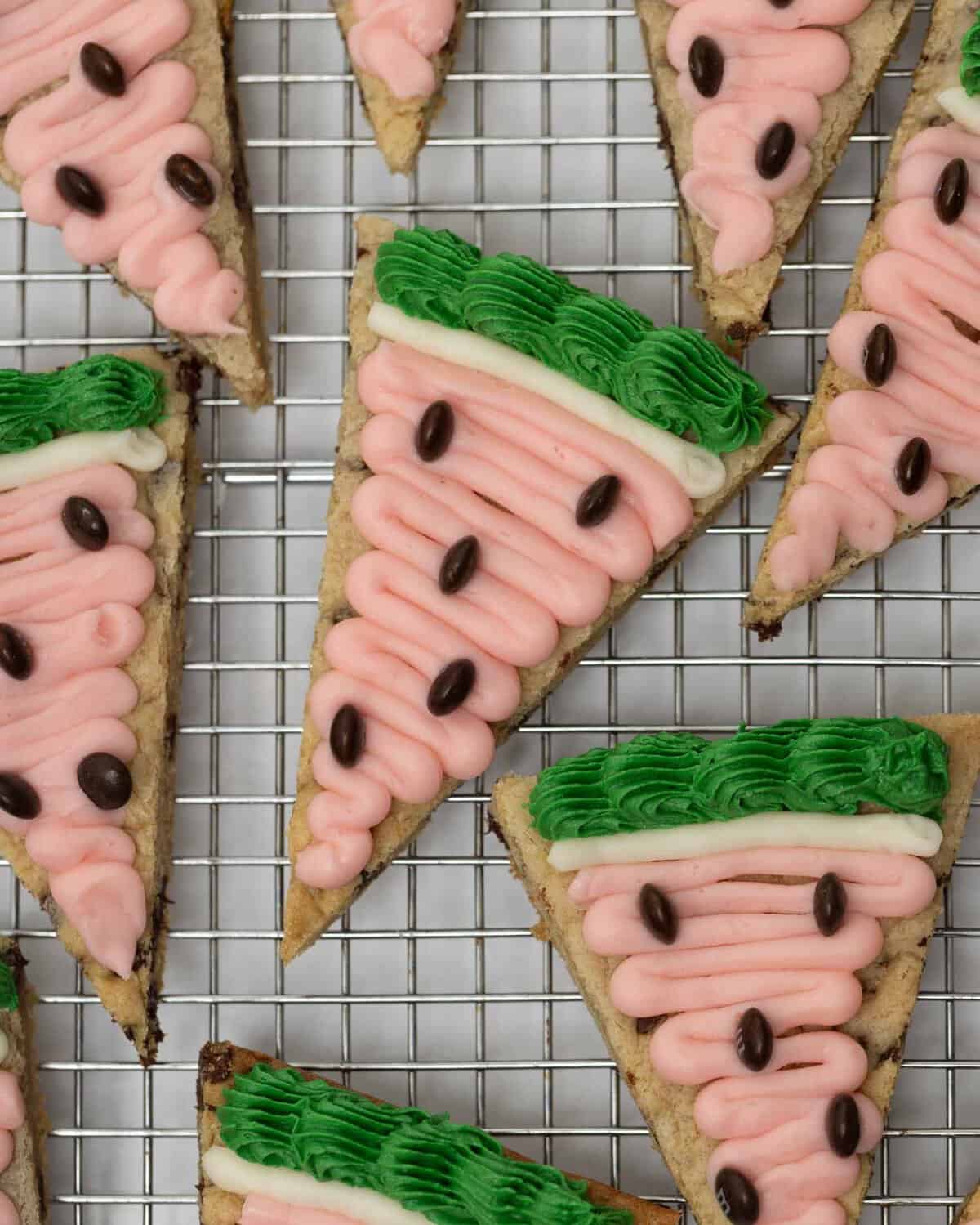Watermelon cookie cake slices with pink frosting, white border, green ruffled rind, and brown M&M seeds on a wire cooling rack