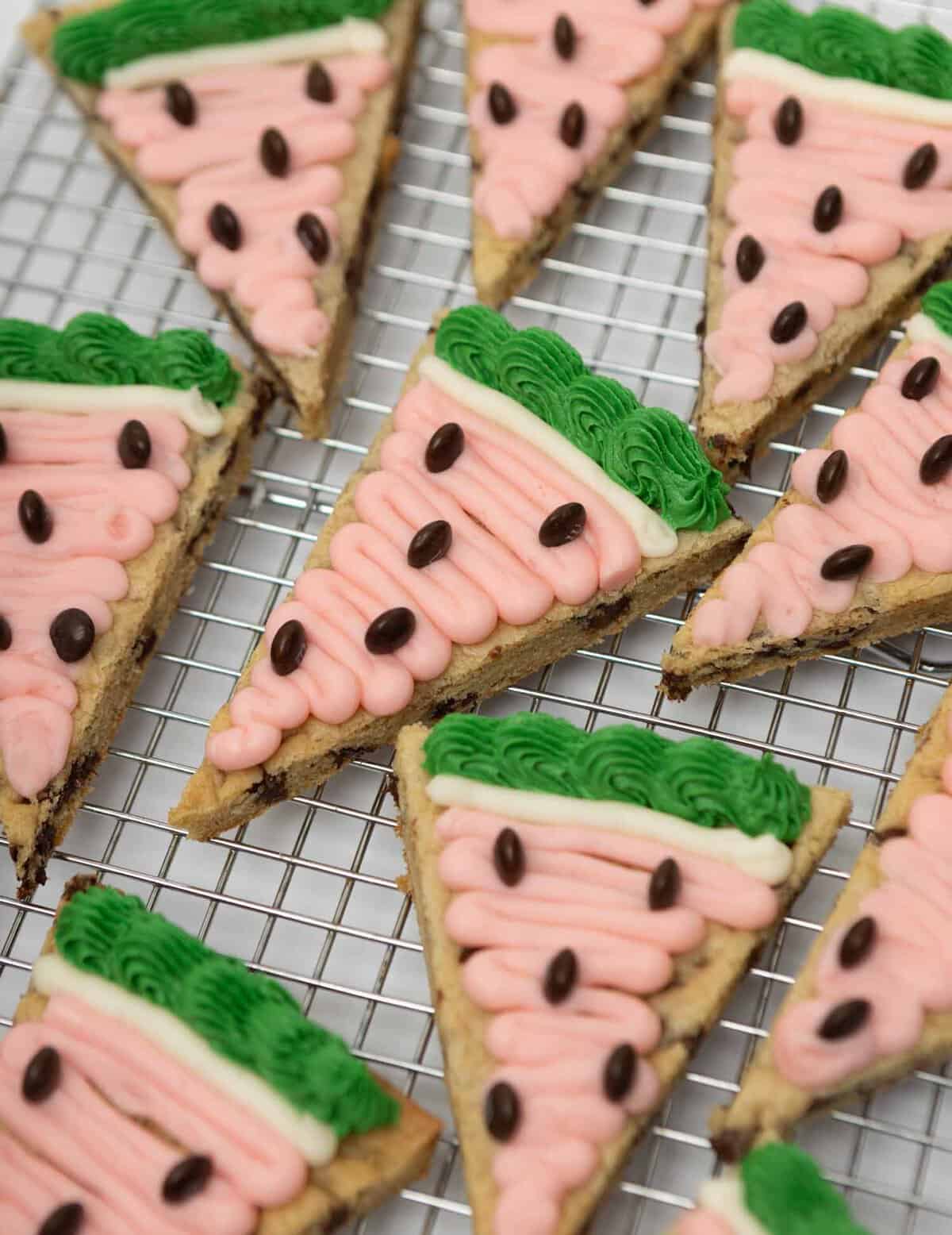 Top-down view of watermelon cookie cake slices on a wire cooling rack, decorated with pink frosting, white ring, green ruffled rind, and brown M&M seeds