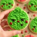 Fuzzy green Grinch Christmas cookie with candy eyes and red cheeks held over a batch of cookies