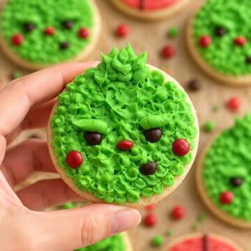 Fuzzy green Grinch Christmas cookie with candy eyes and red cheeks held over a batch of cookies