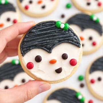 Penguin Christmas cookie with black buttercream, red cheeks and an orange nose held over a batch of cookies on a white background