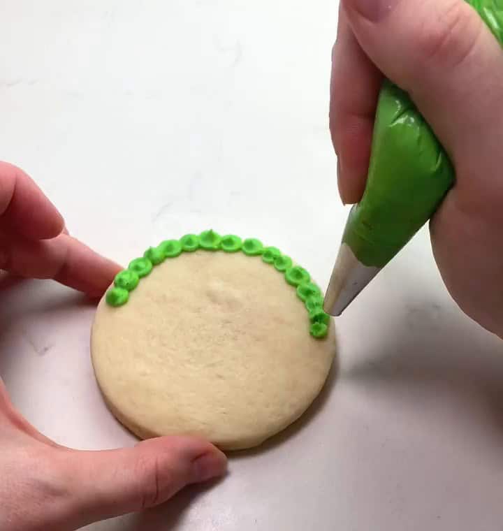 Piping green buttercream fur around the edge of a round sugar cookie