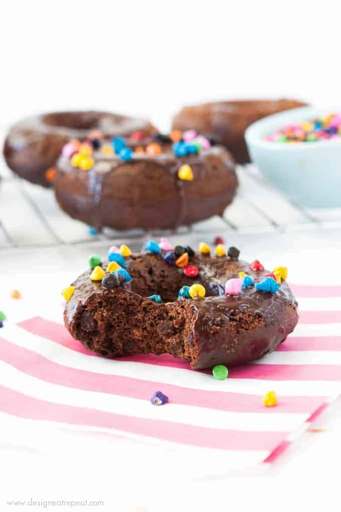 Cosmic Triple Chocolate Cake Donuts on pink striped napkin with rainbow sprinkles and a bite taken out.