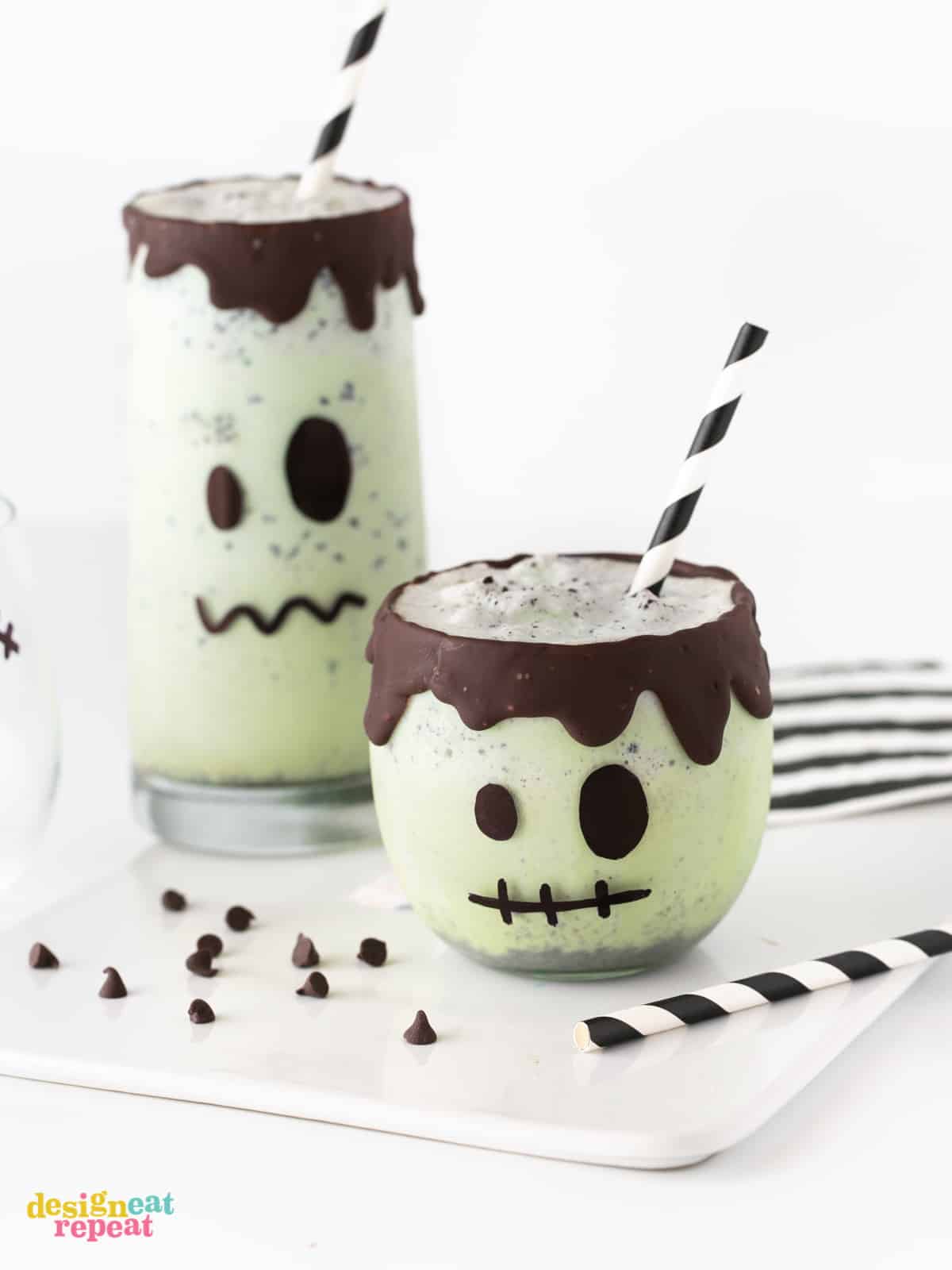 Small mint milkshake decorated like Frankenstein with chocolate rim. With paper black straw and tall Frankenstein green milkshake on white cutting board. Easy Halloween Milkshakes.