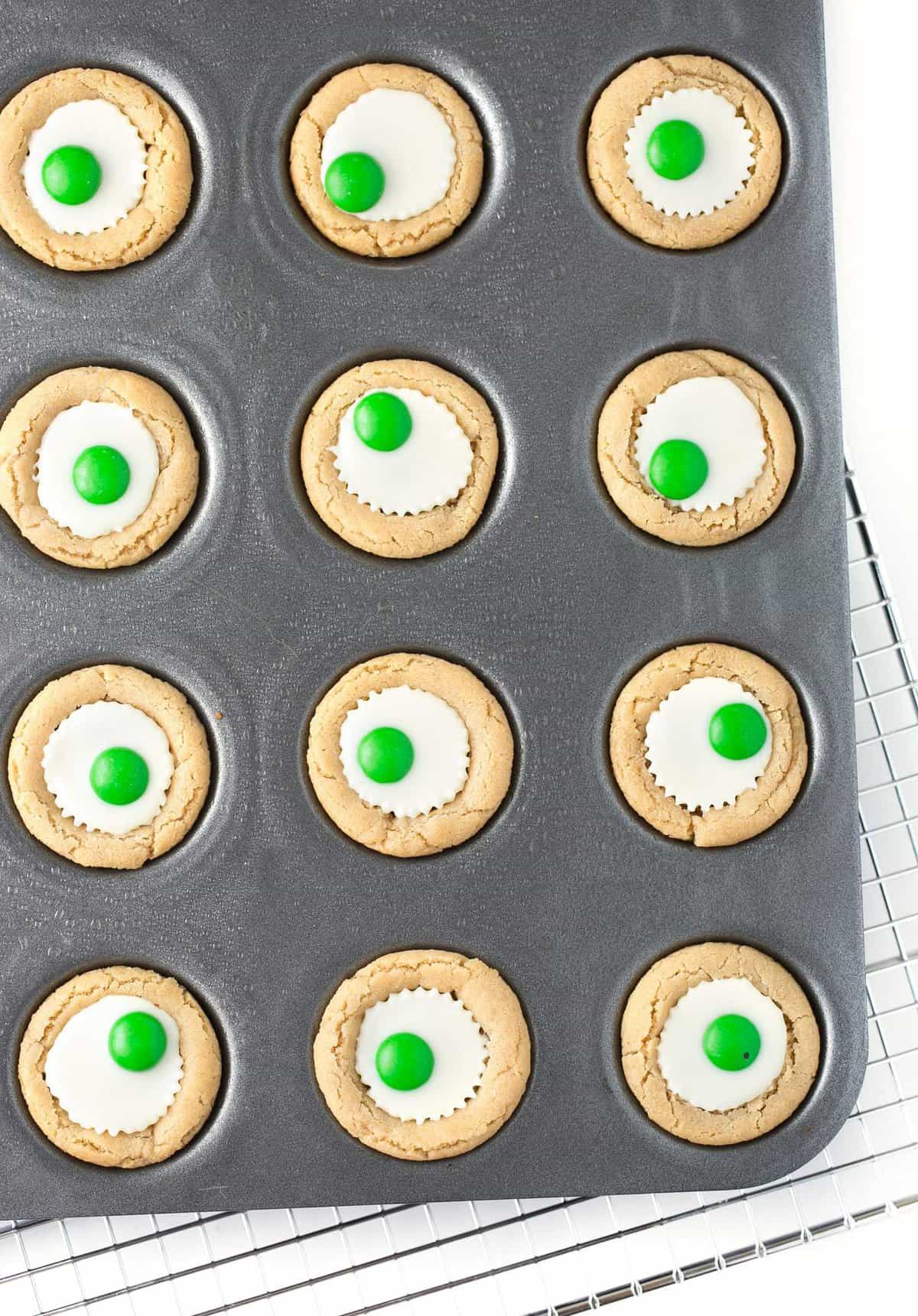 Overhead photo of mini muffin tin with white chocolate peanut butter cup cookies with green M&M's in the center to look like Halloween eyeballs.