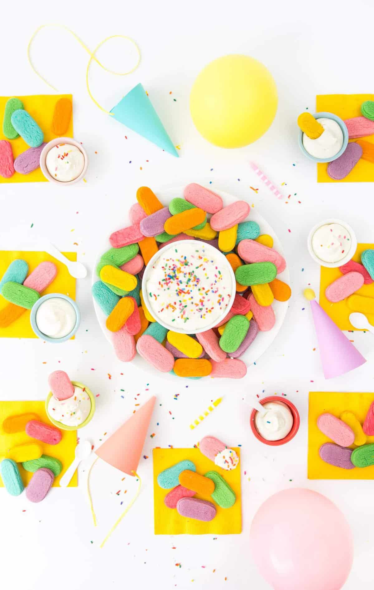 Overhead photo of party table with plate of rainbow sugar cookies on yellow napkins, balloons, and paper party hats.