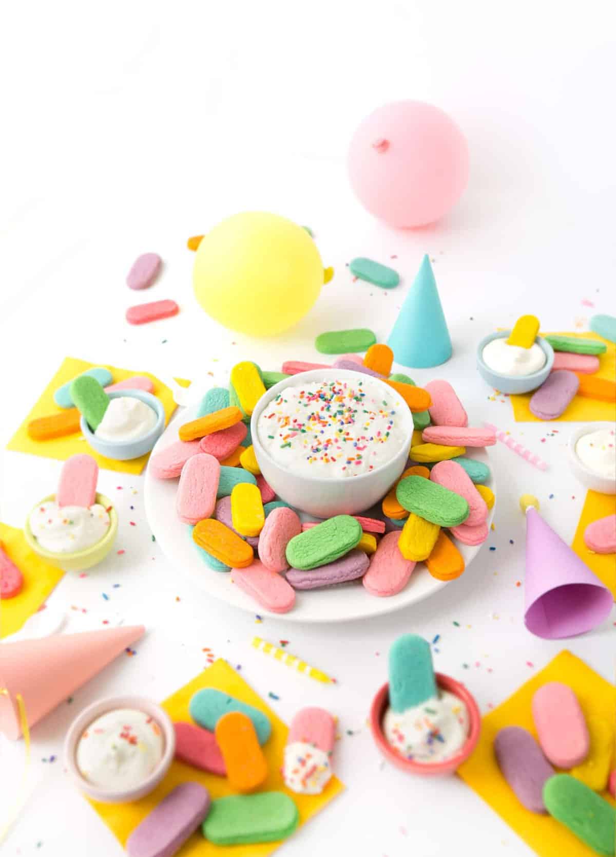 Plate of rainbow sprinkle sugar cookies with a bowl of cream cheese frosting in a party scene.