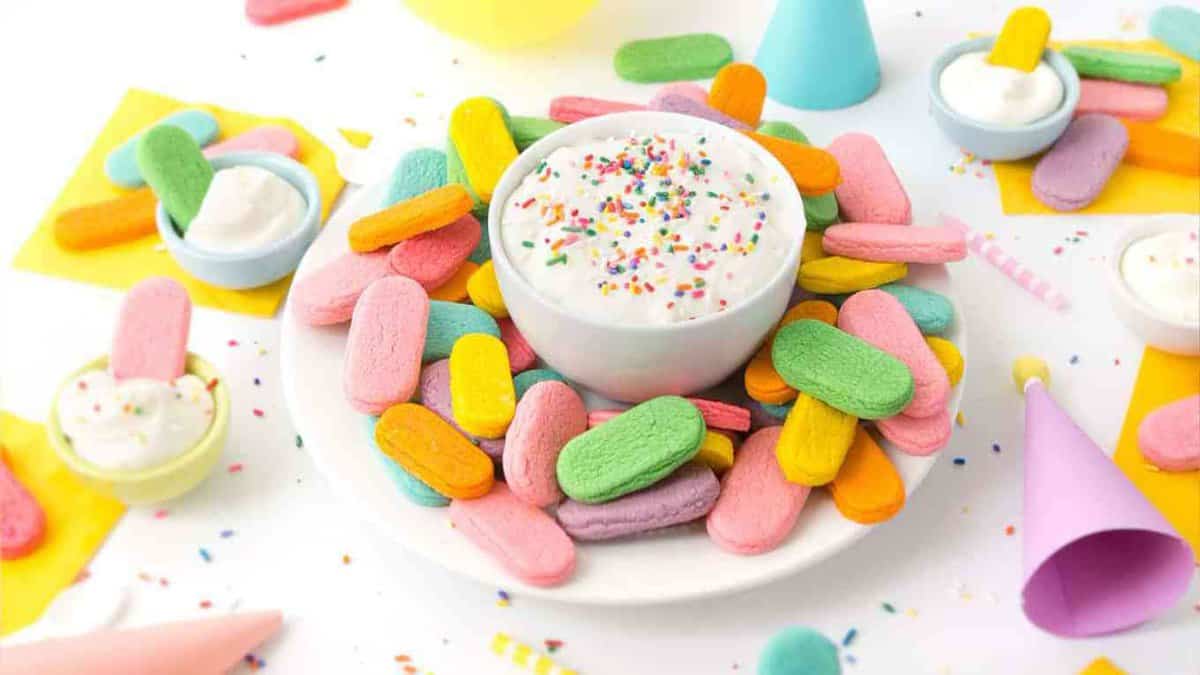 Plate of rainbow sprinkle sugar cookies with a bowl of cream cheese frosting in a party scene.