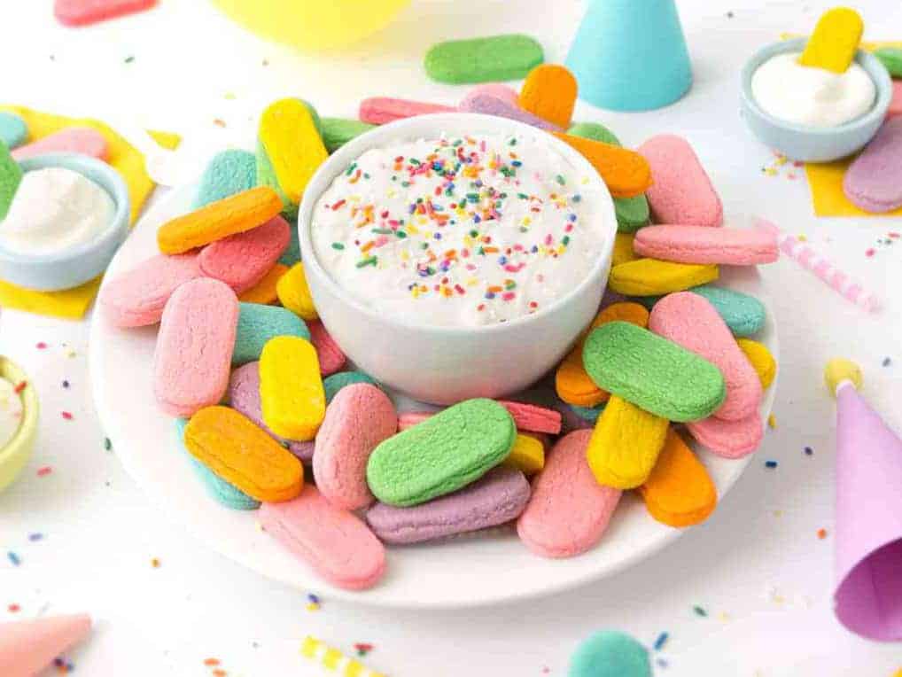 Plate of rainbow sprinkle sugar cookies with a bowl of cream cheese frosting in a party scene.