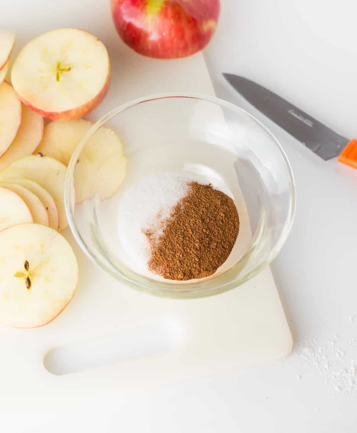 Glass bowl of cinnamon and sugar for apple chips with knife and apple slices.