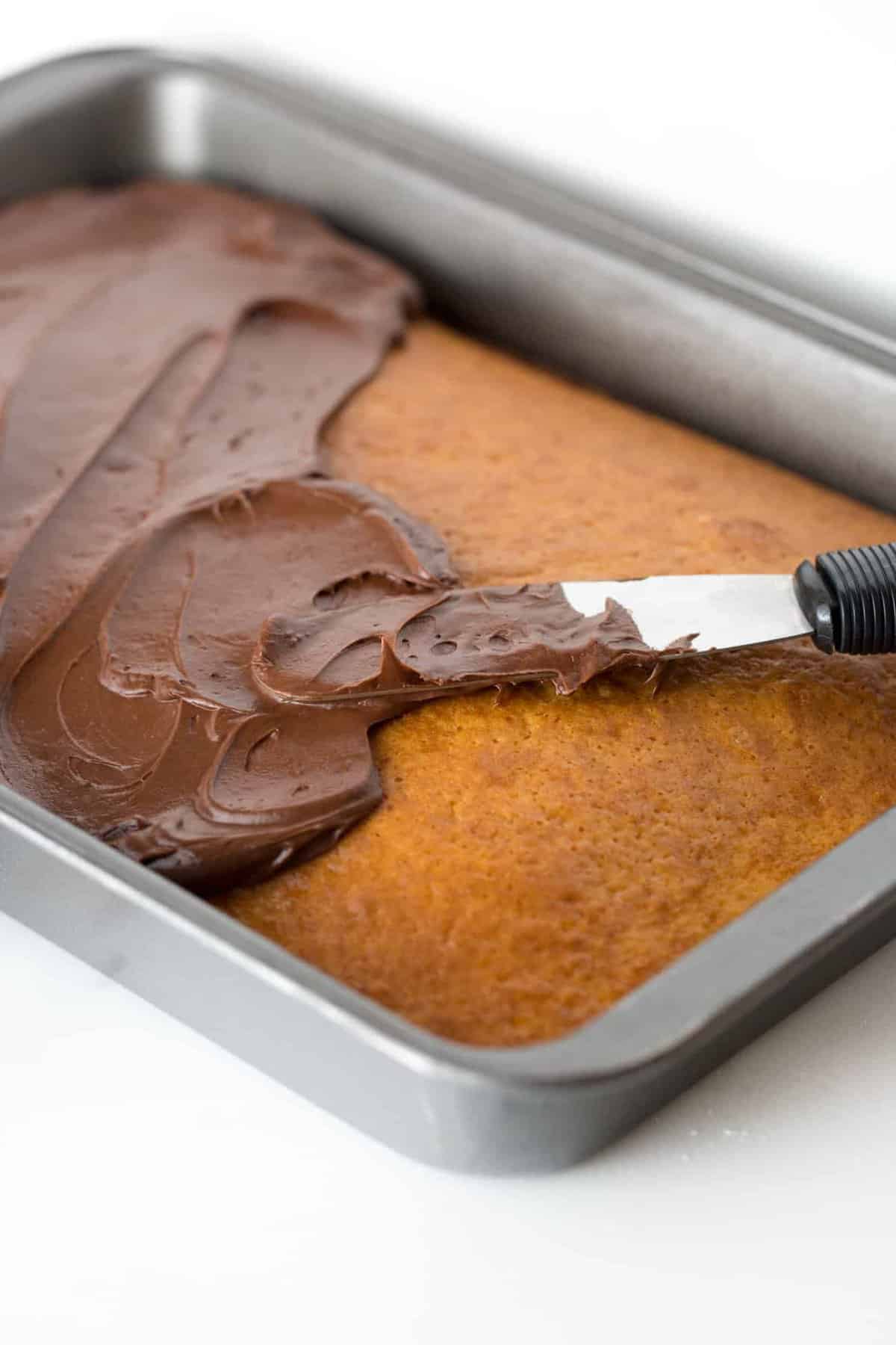 9x13 metal cake pan with yellow cake, spreading easy chocolate frosting on top with cake spreader.