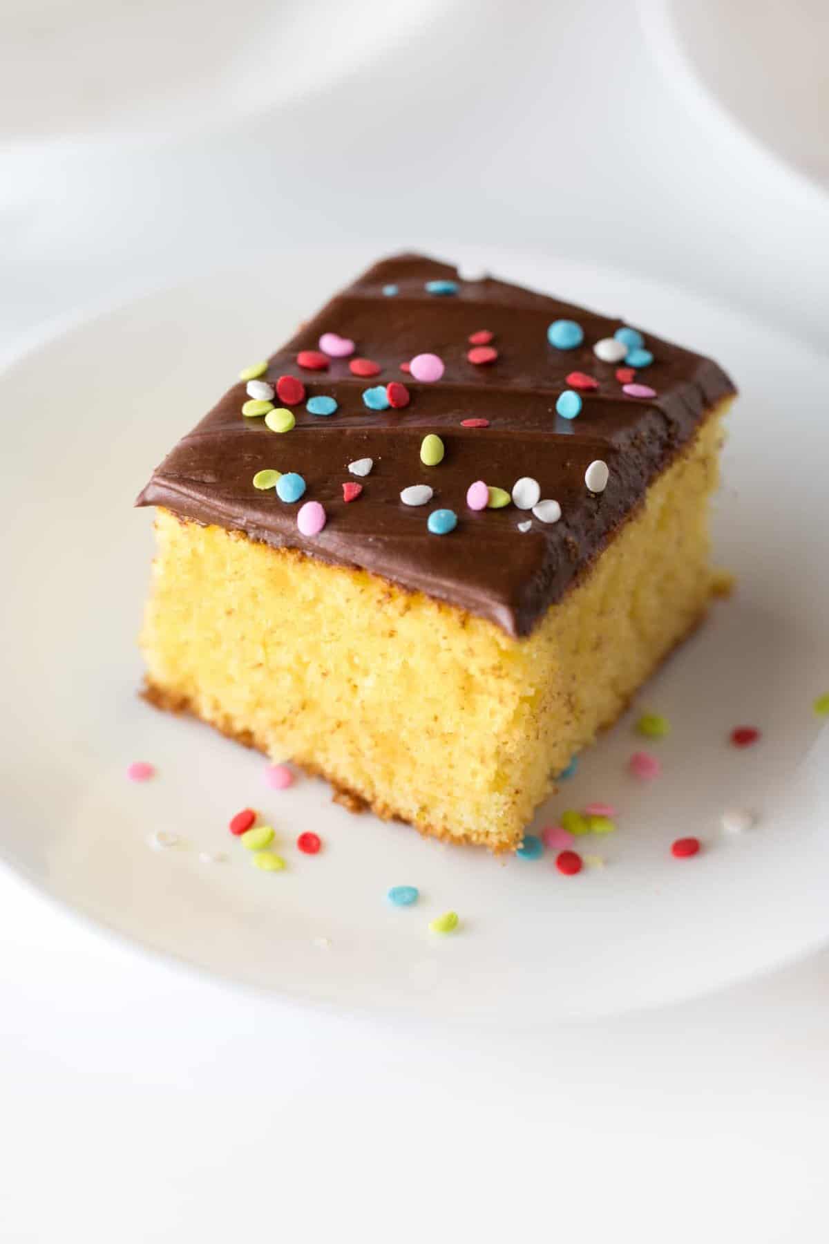 Square slice of yellow cake with easy chocolate frosting with rainbow sprinkle quins on top. On small white plate.